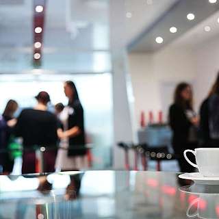Tisch mit einer Kaffeetasse im Vordergrund. Im Hintergrund stehen Menschen und unterhalten sich.
